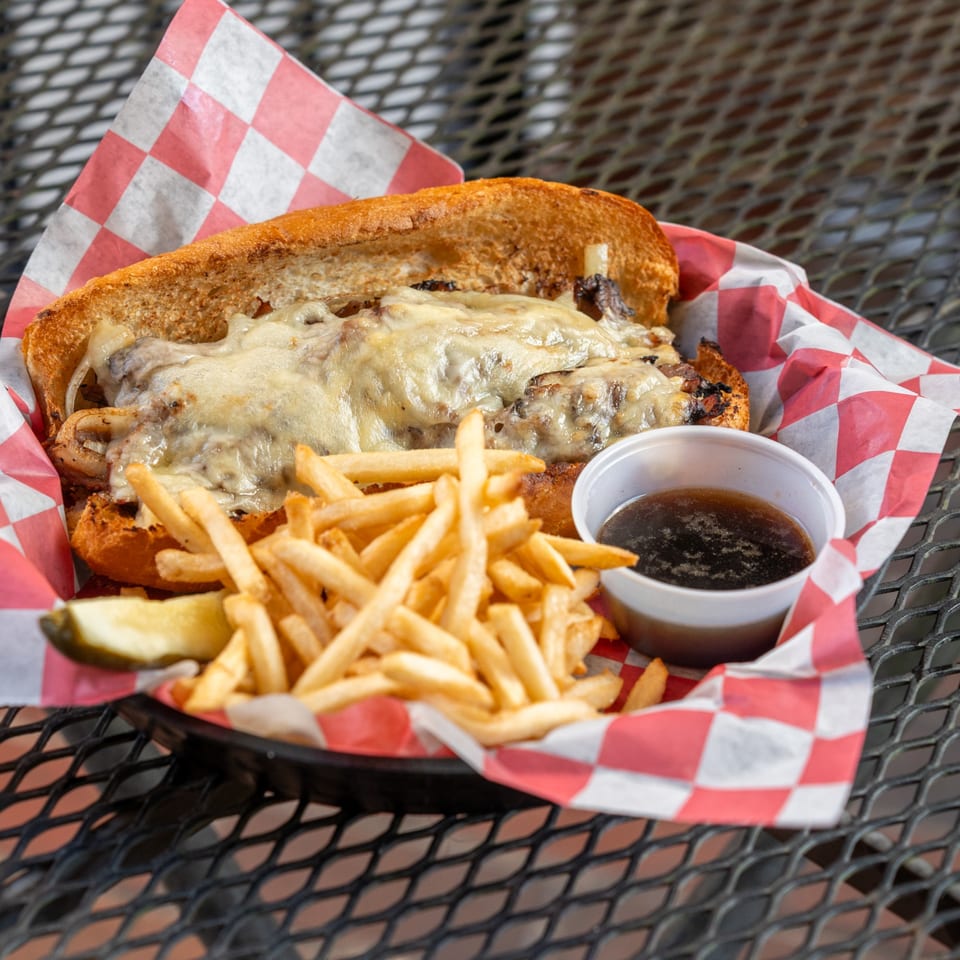 Cheesesteak sandwich with melted cheese, fries, and a side of au jus in a red checkered basket.